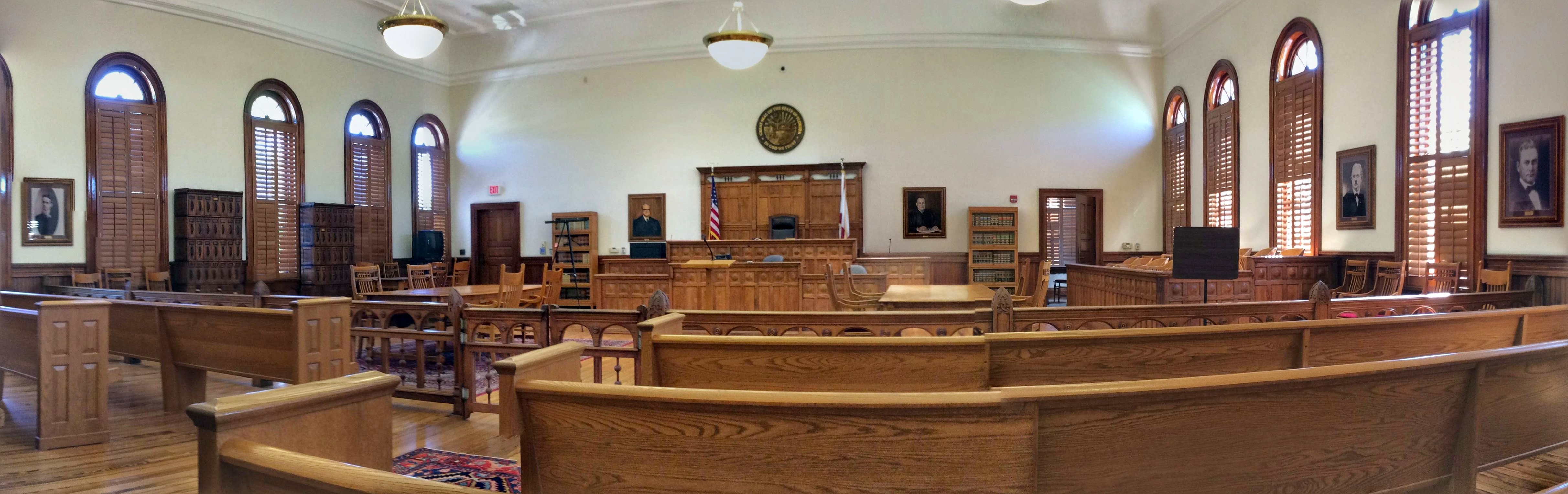 Panoramic interior of the Nassau County, Florida courtroom: dark wood pews, judge's bench, jury box, arched windows with shutters, Florida state seal above the bench